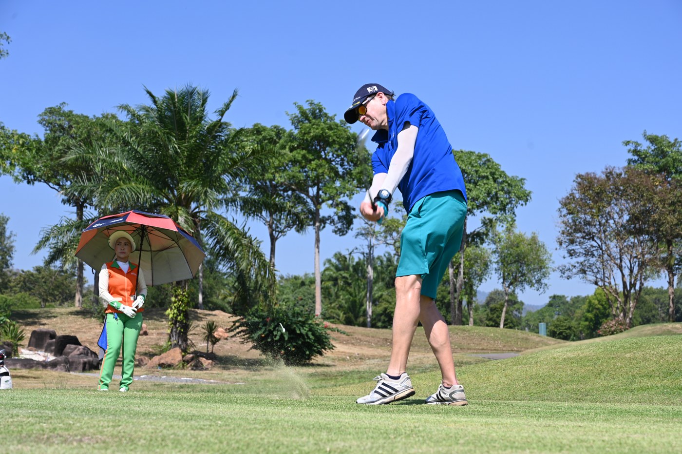 A golfer swings while a caddie waits under a sun umbrella on a real course